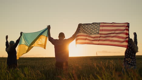 Young people raise up the American and Ukrainian flags against the background of a wheat field where the sun rises