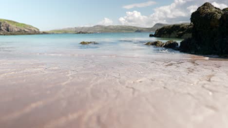 Nahaufnahme-Mit-Geringer-Schärfentiefe-Des-Wassers,-Das-Stetig-über-Einen-Strand-Und-In-Ein-Türkisfarbenes-Meer-Fließt-Und-Den-Sand-Erodiert,-Während-Im-Hintergrund-Langsam-Wellen-Brechen