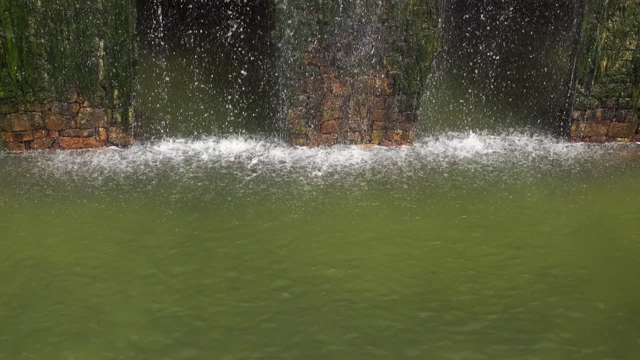 The water flows abundantly from the waterfall over the brick wall covered with green algae. The green color is visible in the water's depth, giving a strong sense of dimension to this scene
