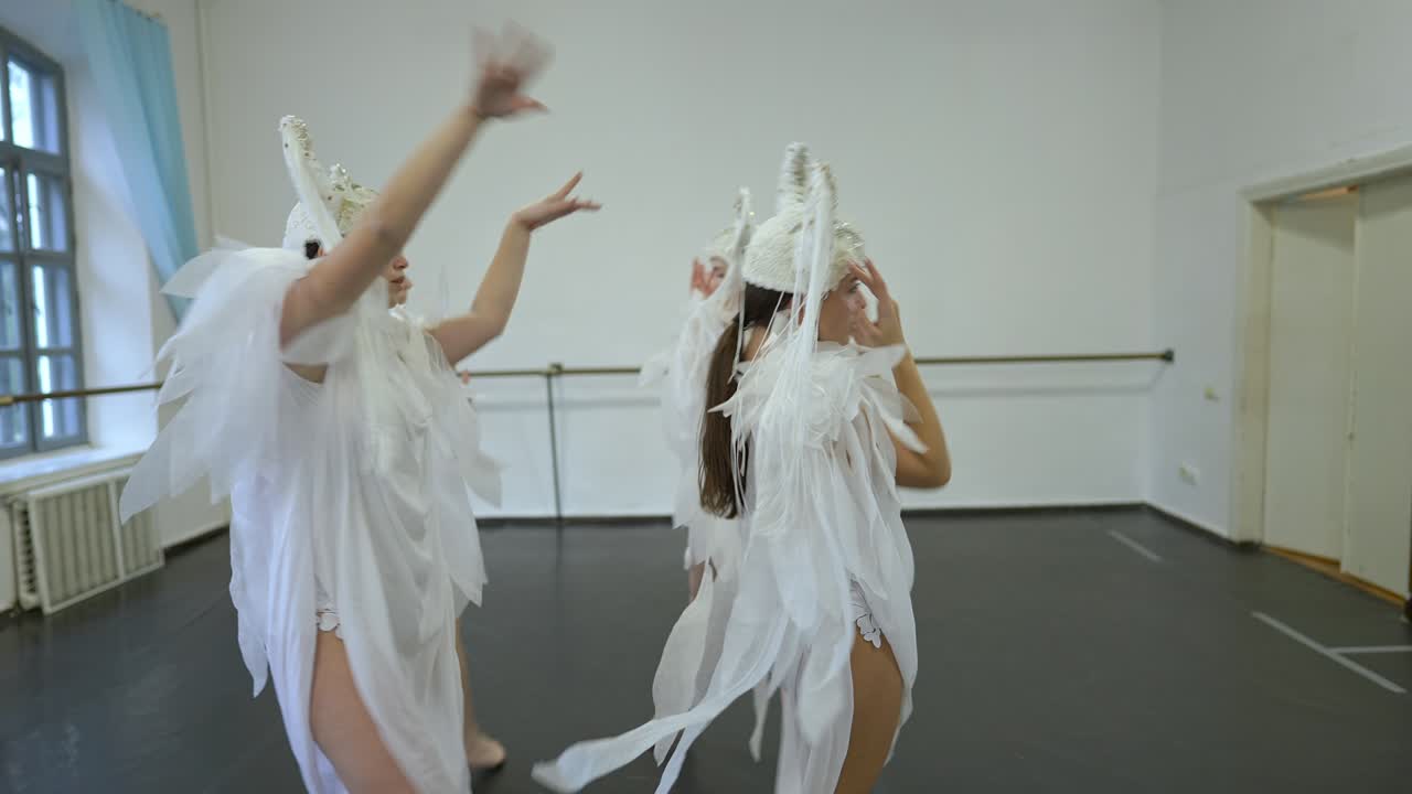 Graceful talented young Caucasian women walking in circle in slow motion rehearsing dance in studio. Beautiful slim dancers in white angel costumes dancing indoors moving hands.