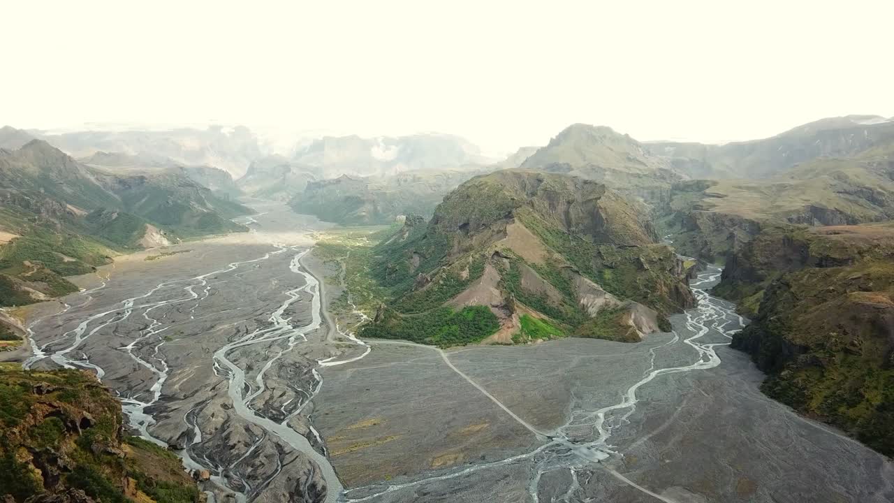 Thórsmörk, Iceland, features a vast braided river system surrounded by rugged green and brown mountains under a soft light. This dramatic landscape highlights the raw beauty of Iceland's nature