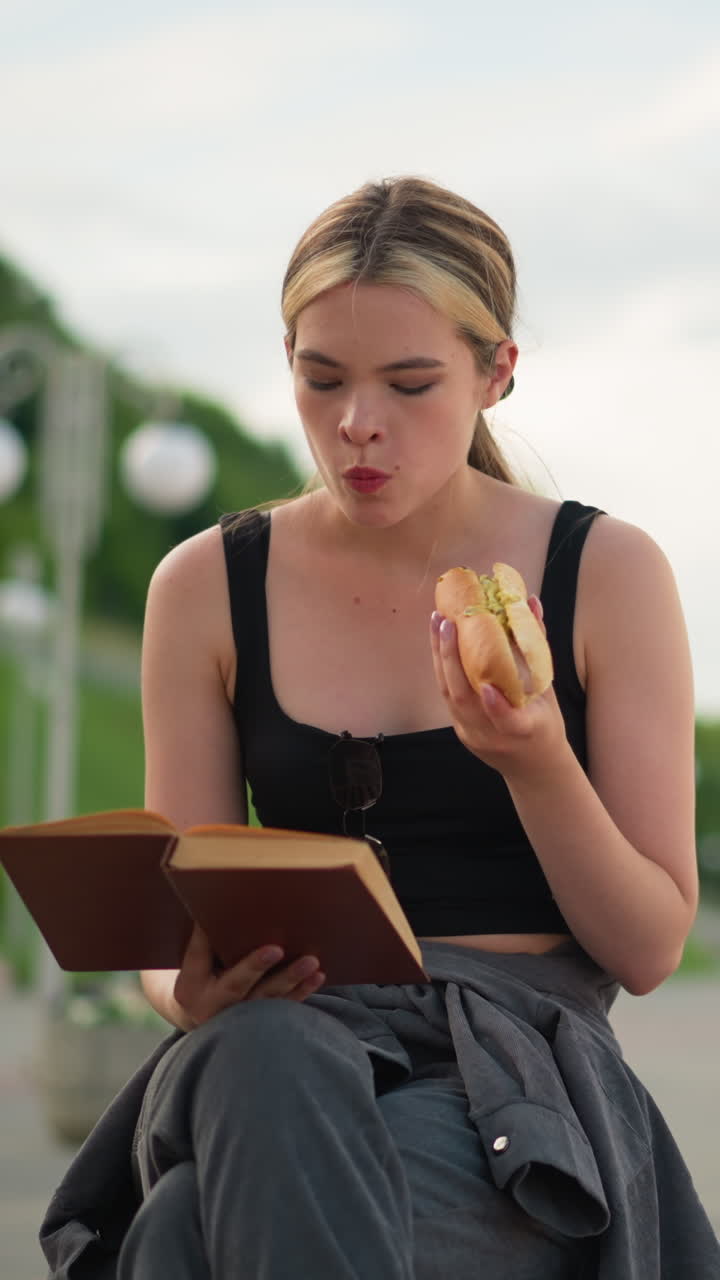 mujer sentada al aire libre con las piernas cruzadas sosteniendo un libro en una mano mientras come una hamburguesa y lee, el fondo presenta un poste de lámpara con un ambiente al aire libre sereno y borroso