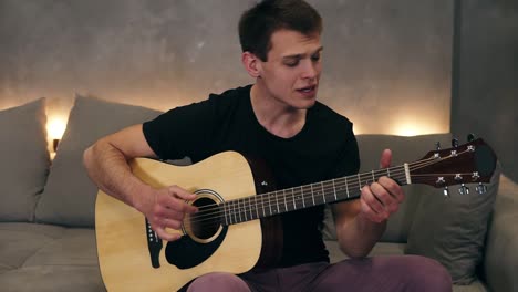 Close-up-portrait-of-a-cheerful-caucasian-man-singing-a-song.-Playing-on-an-acoustic-guitar-and-enjoying-it,-while-sitting-on-a-grey-couch-with-a-backlight-under