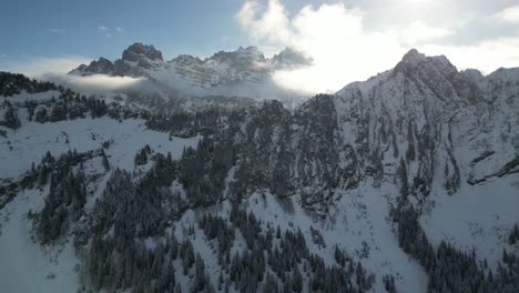 Umlaufbahn-Parallaxe-Um-Schneebedeckten-Wald-Im-Sattel-Mit-Gezackten-Scharfen-Bergrücken-Im-Hintergrund,-Während-Wolken-Das-Sonnenlicht-Streuen