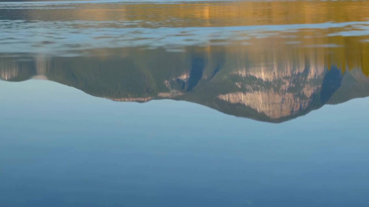 Mirror-like mountain ridge and golden treeline reflect on calm blue lake surface that develops ripples and low waves across narrow reflected band under warm directional sunlight, with reflection