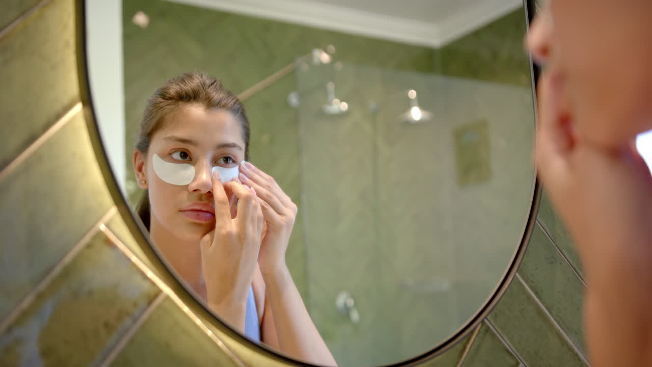 Concentrata ragazza adolescente birazziale che applica maschere contorno occhi guardandosi allo specchio del bagno, rallentatore