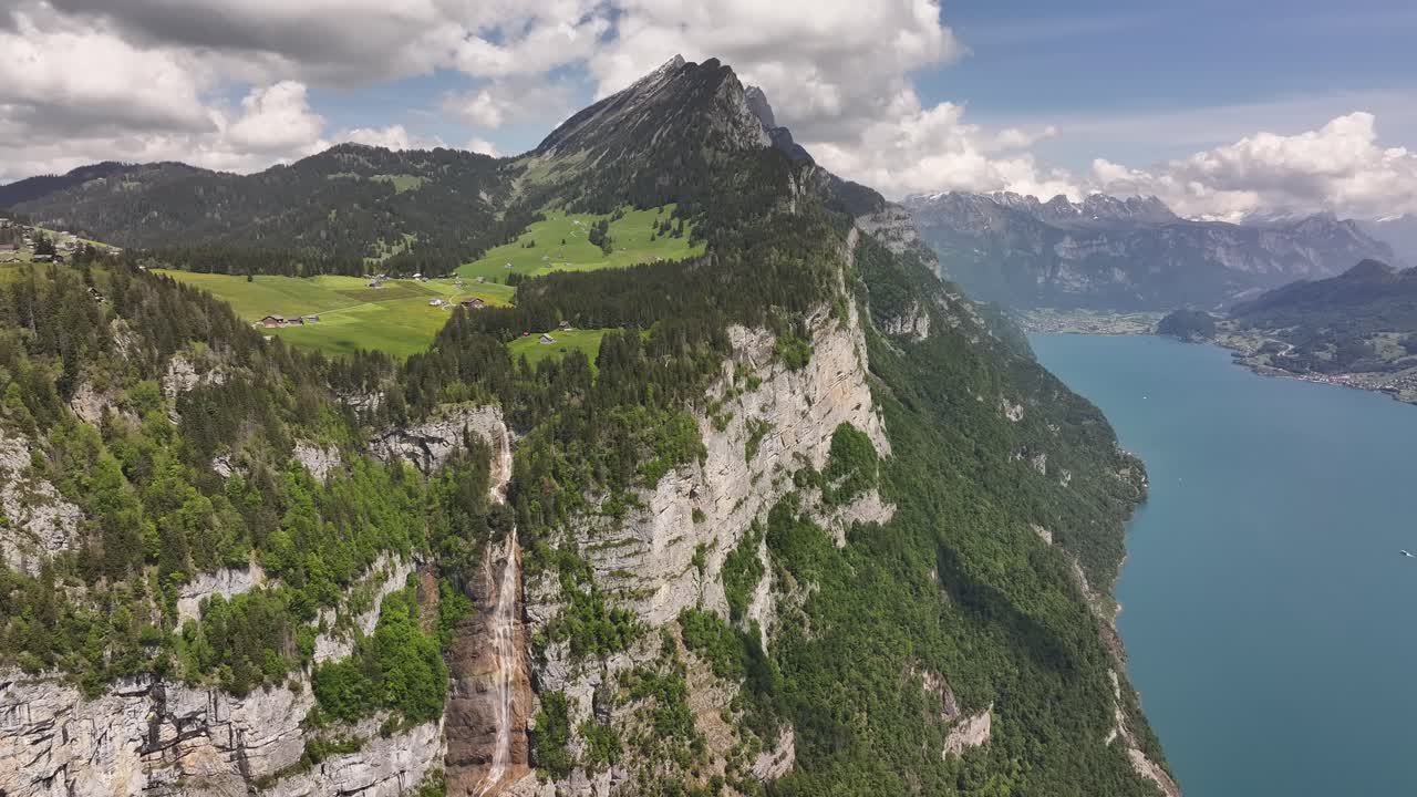 Dramatic aerial of Walensee in Switzerland, showcasing steep cliffs, lush green meadows, alpine peaks, and a cascading waterfall meeting the turquoise lake below