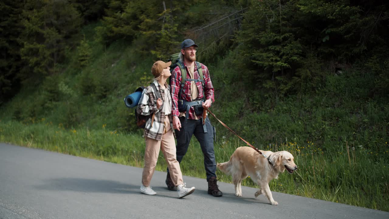un tipo rubio con una camisa roja y una chica rubia con ropa especial para caminar caminan con su perro de color claro a lo largo de la carretera a lo largo del bosque y viajan a través de nuevos territorios