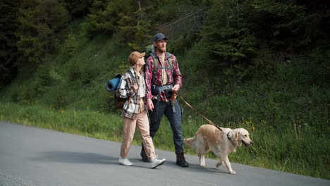 a blond guy in a red shirt and a blond girl in special clothes for hiking walk with their dog light-colored along the road along the forest a they travel through new territories