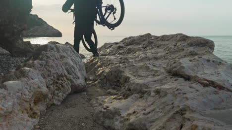 man carefully carries his bike over rocks, showcasing his determination and resilience near sea coast