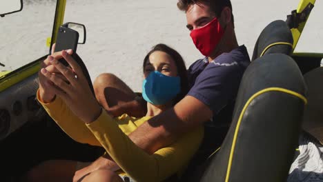 Happy-caucasian-couple-wearing-face-masks-sitting-in-beach-buggy-taking-selfies