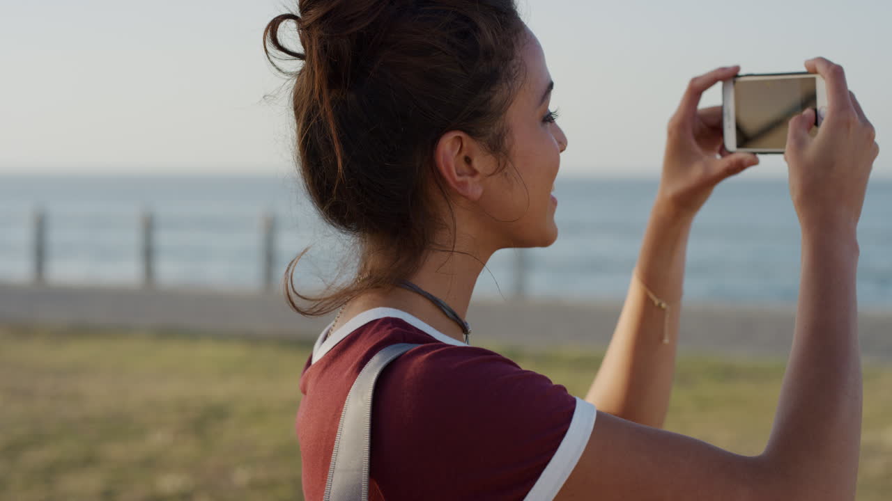 초상화 매력적인 젊은 히스패닉 여성 관광객이 스마트폰을 사용하여 사진을 찍고 바다 해변에서 아름다운  ⁇ 은 휴가를 즐기고 경험을 공유합니다.