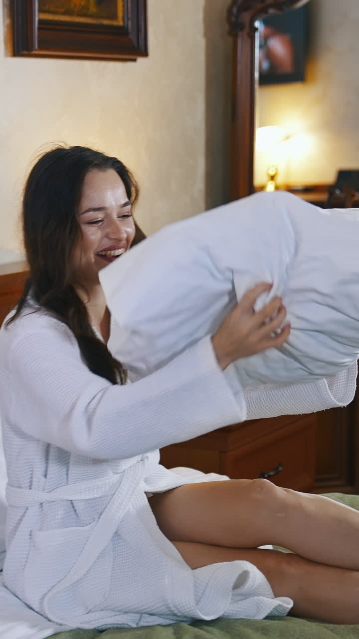 Beautiful woman on bed in hotel. Happy young woman in white bathrobe sitting on bed and throwing up pillow. Pleasant vacation. Vertical video