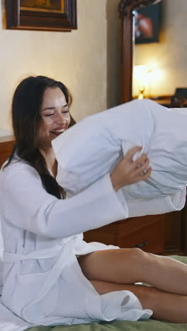 beautiful woman on bed in hotel. happy young woman in white bathrobe sitting on bed and throwing up pillow. pleasant vacation. vertical video