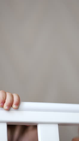 cheerful happy baby boy playing hide-and-seek in his crib. lovely laughing baby sits and stands up in white cot. close up. vertical video