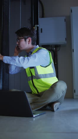 video vertical de un hombre asiático en chaleco de alta visibilidad usando una computadora portátil hablando por teléfono en la sala del servidor