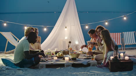 Grupo-De-Amigos-Cenando-Sentados-En-La-Playa,-Con-Una-Mesa-Baja-Y-Una-Carpa