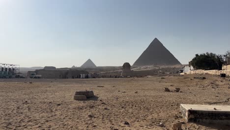 Wide-shot-of-the-Giza-pyramid-complex-under-clear-skies-at-daytime,-sandy-foreground-with-sparse-visitors