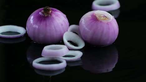 Sliced-raw-onion-rings-falling-onto-whole-and-chopped-onions-on-rippling-water-surface-isolated-on-black-background