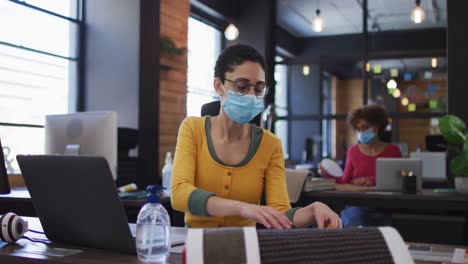 mujer caucásica con máscara de cara leyendo documentos y usando una computadora portátil en una oficina moderna