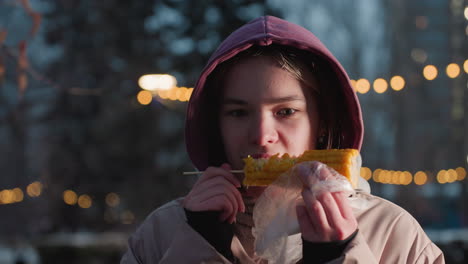 joven comiendo maíz en un palo envuelto en plástico blanco con luces bokeh en un fondo borroso, vapor que se eleva desde el maíz fresco, disfrutando de un bocadillo en un entorno al aire libre durante el invierno