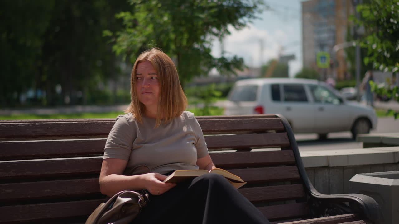 Woman sitting on wooden bench outdoors holding open book, gazing away with thoughtful expression under sunlight, surrounded by greenery and urban background