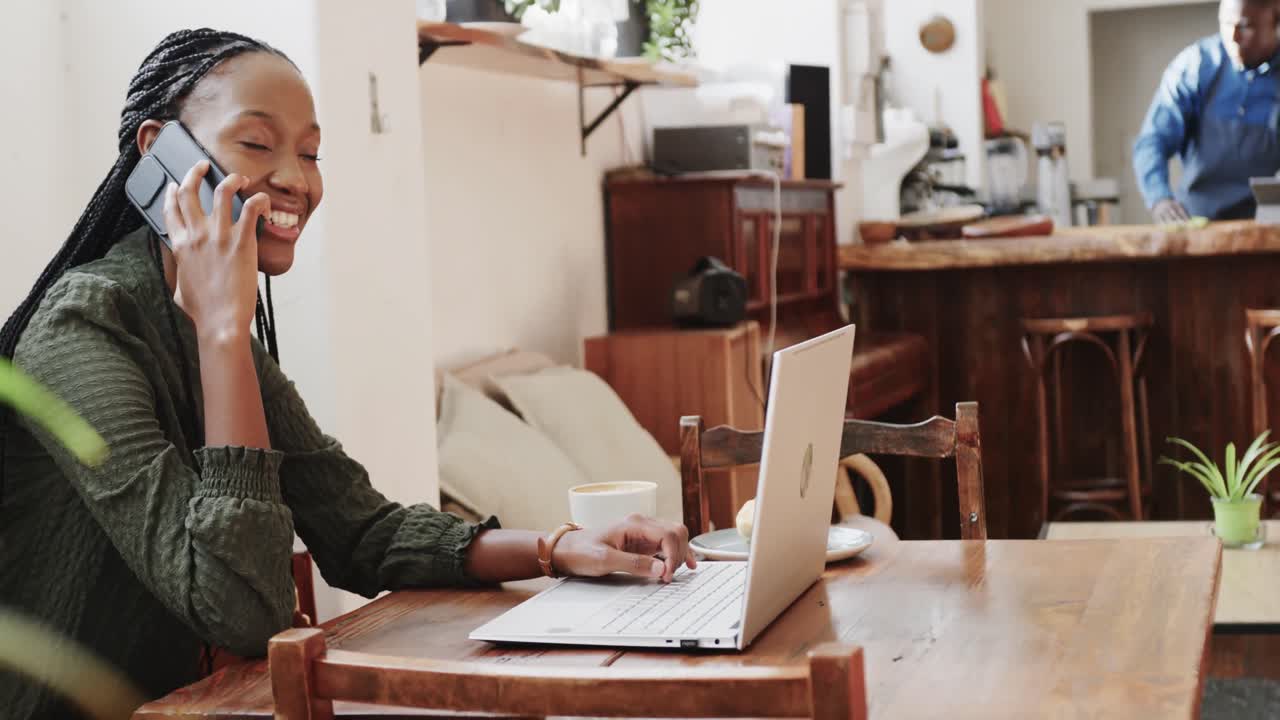 Heureuse femme afro-américaine utilisant un ordinateur portable et parlant au smartphone dans un café, au ralenti.