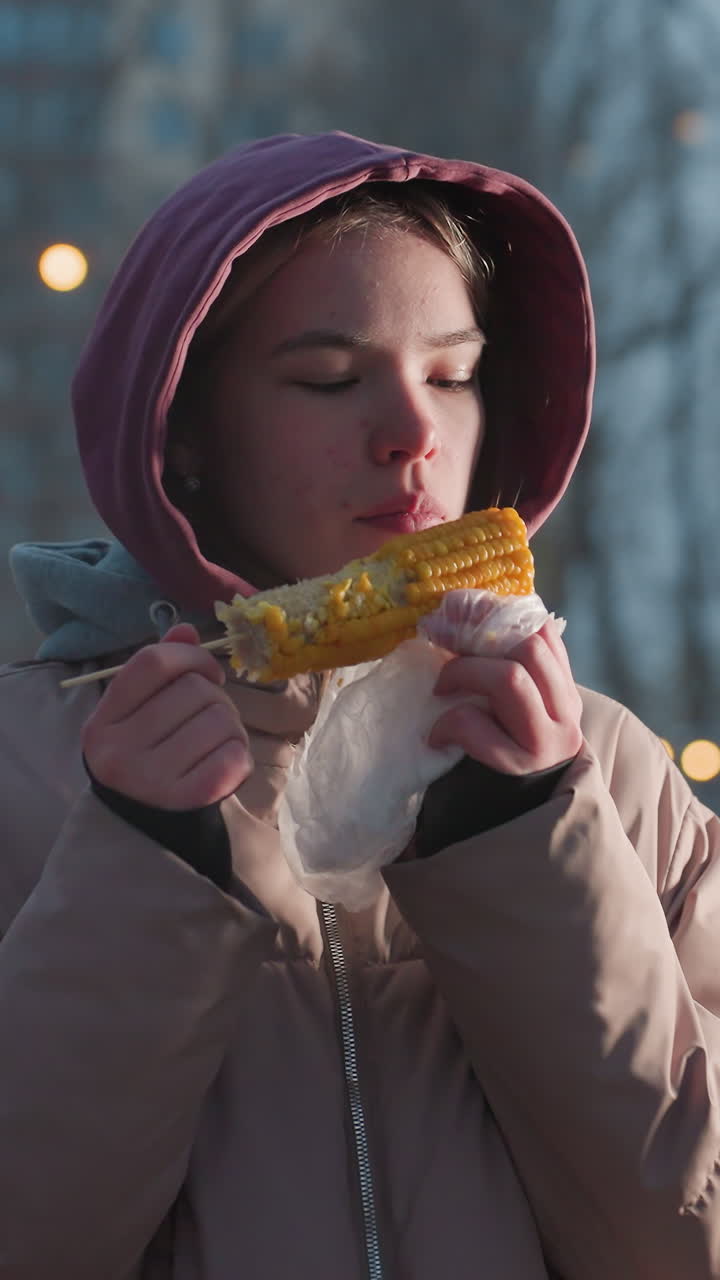 joven disfrutando de maíz en un palo envuelto en una bolsa blanca con luces bokeh en un fondo borroso, vapor que se eleva desde el maíz fresco, personas caminando en el fondo durante el tiempo de bocadillo al aire libre de invierno