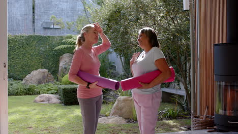 holding yoga mats, two multiracial female friends chatting and smiling in outdoor garden setting
