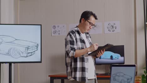 vista lateral de un hombre asiático dibujando un nuevo concepto de diseño de automóvil en una tableta mientras camina por el estudio con tv y computadoras muestra modelo de automóvil eléctrico 3d