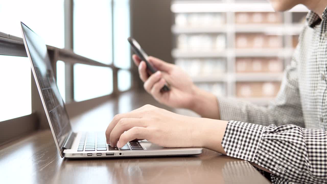 Giovane uomo d'affari asiatico che usa un computer portatile in uno spazio di lavoro con smartphone e taccuino su una scrivania di legno. Mano maschile che digita sulla tastiera del portatile. Concetto di stile di vita freelance nell'era digitale