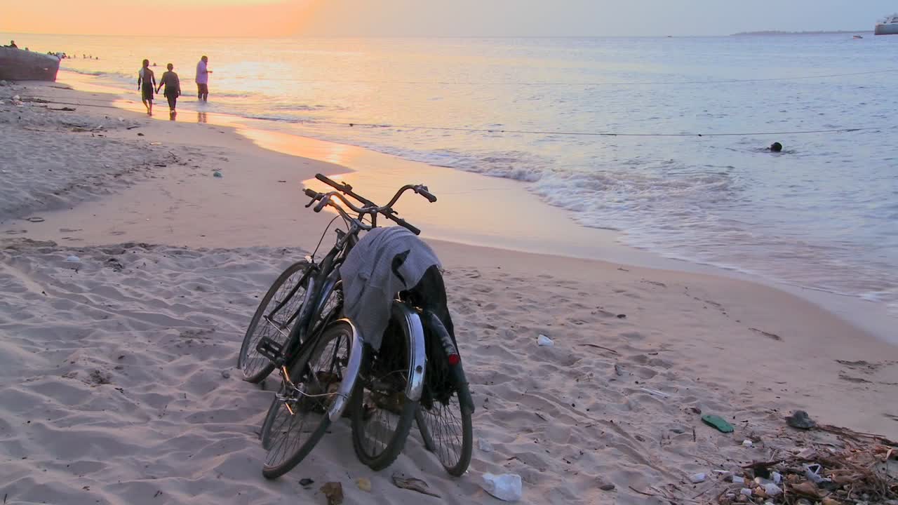두 대의 자전거가 해안에 주차되어 있고 아이들이 멀리서 놀고 있는 해변을 따라 일몰이 촬영되었습니다.