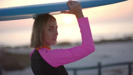 young caucasian woman carries a surfboard at sunset