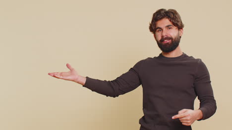 lebanese young man showing thumbs up and pointing at empty blank place against beige background
