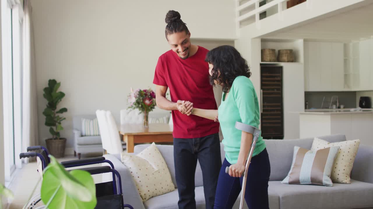 hombre biracial sonriente con dreadlocks ayudando a su pareja a la silla de ruedas en la sala de estar