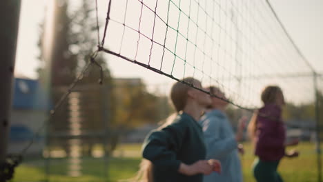 slightly blurred women in hoodies engage in volleyball training outdoors near net with golden sunlight glow as they jump with hands raised, background features greenery, and building