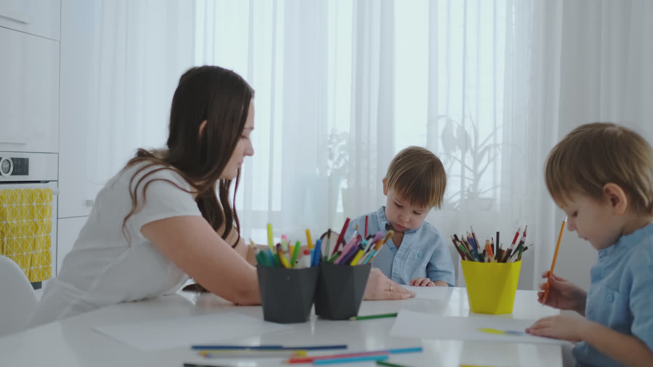 mamá ayuda a los hijos a aprender a dibujar haciendo la tarea de preparación preescolar en casa sentado en la cocina blanca.