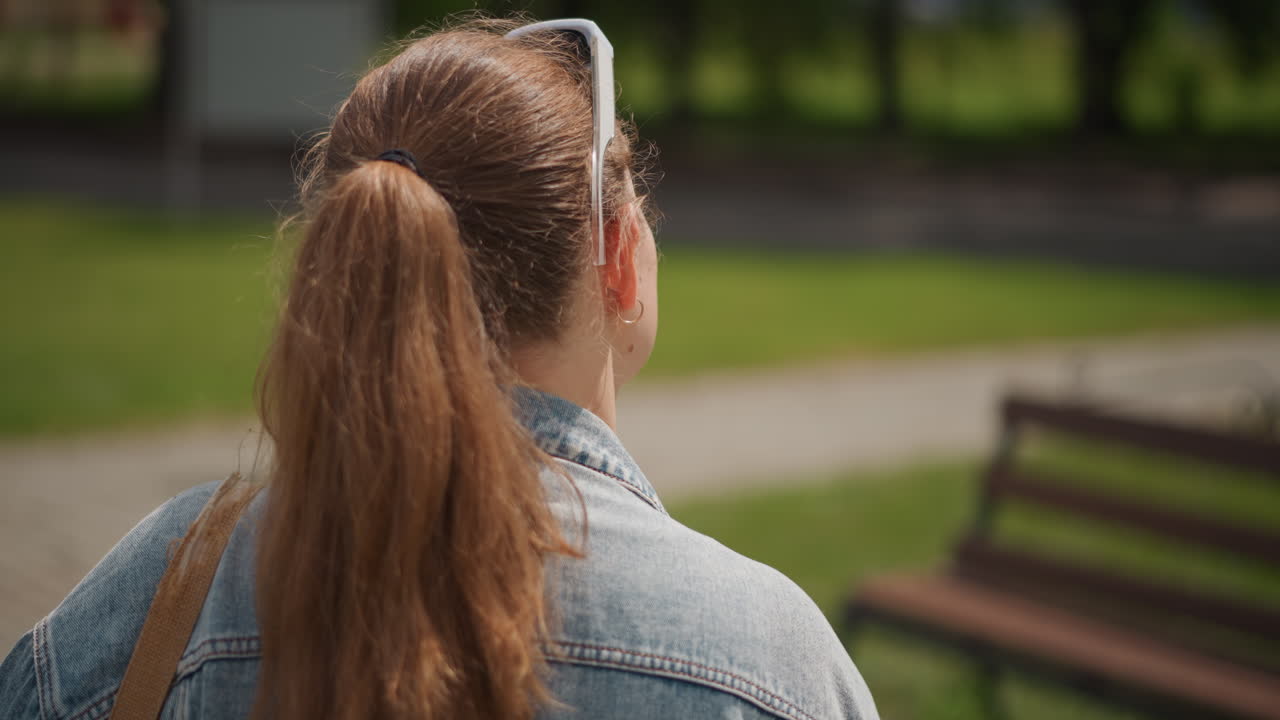 Kvinde med hestehale på solrig parkbænk. Ung kvinde sidder på parkbænk og nyder sollys. Teenager kvinde med hestehale sidder på bænk midt i frodigt grønt landskab.