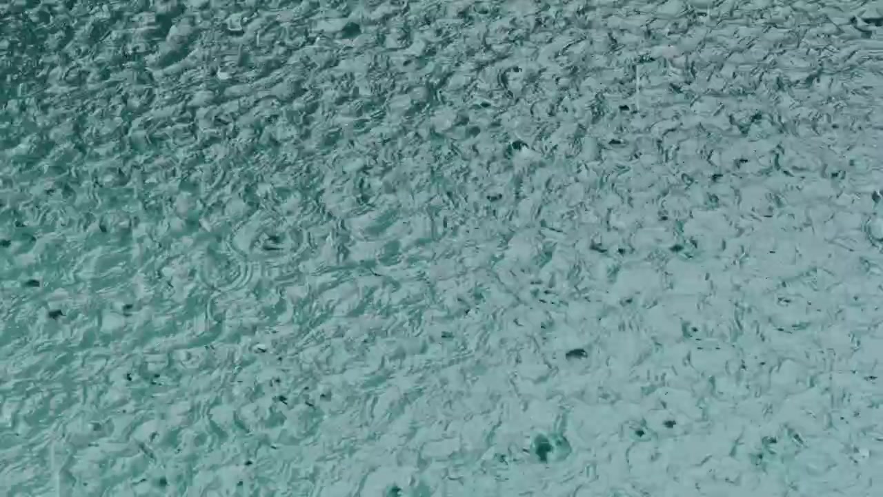 Close-up of raindrops falling into turquoise blue water, forming beautiful circular ripples, abstract patterns, and calming motion.