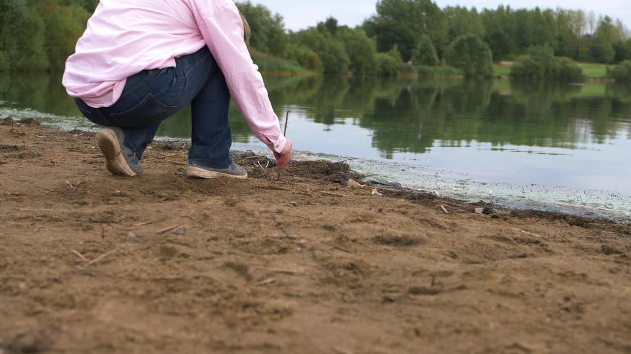 persona solitaria garabateando en arena dorada con un palo junto al lago, una amplia toma de paisaje