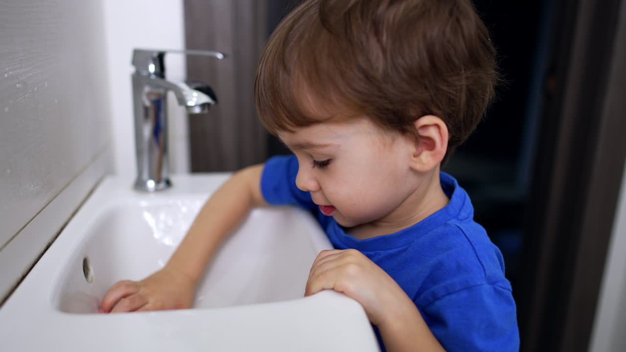Adorable Caucasian kid stands in the bathroom near the sink. Cute child playing with a drain. Close up.