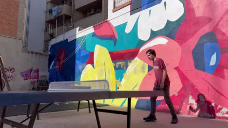 muchacho de 20 años jugando al ping-pong en una mesa de tenis de la calle con su padre con un graffiti de flores de colores en la espalda