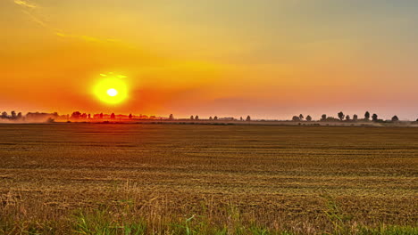 Time-lapse-of-vibrant-sunset-above-machinery-harvesting-on-countryside-farmlands