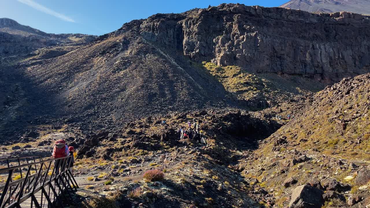 Group of hikers cross a bridge and hike rocky pathway toward mountain bluff at Tongariro Alpine Crossing, New Zealand