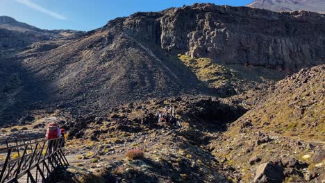 group of hikers cross a bridge and hike rocky pathway toward mountain bluff at tongariro alpine crossing, new zealand
