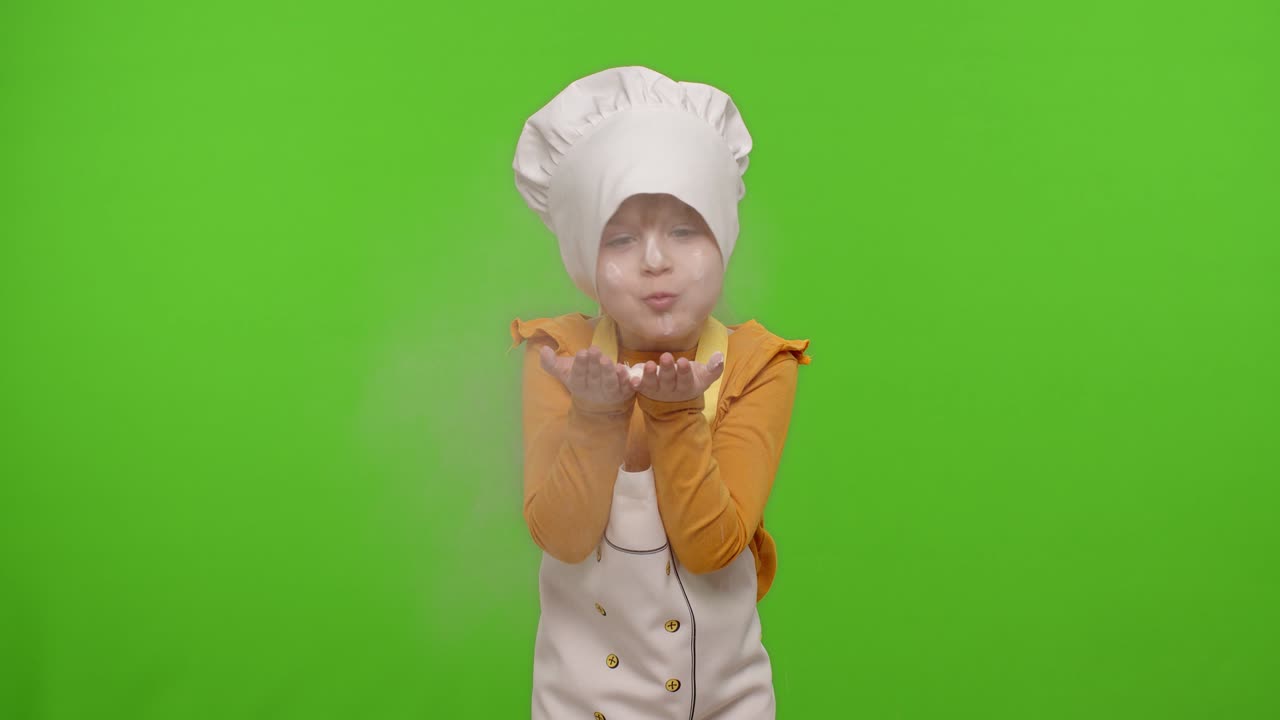 Bambina vestita da cuoca professionista che soffia farina dalle mani verso la telecamera su chroma key.