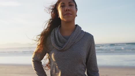 Portrait-of-mixed-race-woman-holding-yoga-mat-at-the-beach-smiling