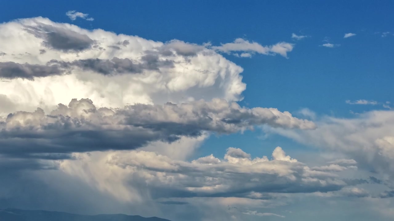4K UHD drone timelapse detailing the dramatic tail end of a monsoon storm cloud. Heavy, layered white and gray clouds shift dynamically against a vivid blue sky, with visible rain falling below.