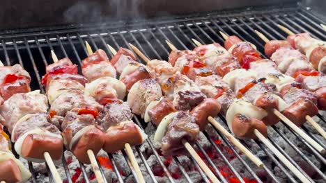 horizontal video panning right to left of meat skewers on a charcoal grill, with red peppers, white onions, and sausages. glowing red coals and light smoke create a rustic, savory scene.