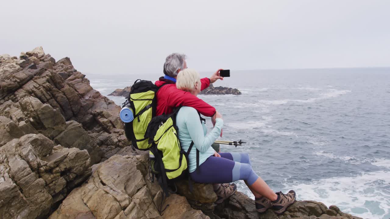 pareja de excursionistas mayores con mochilas sentados en las rocas y tomando una selfie en el teléfono inteligente